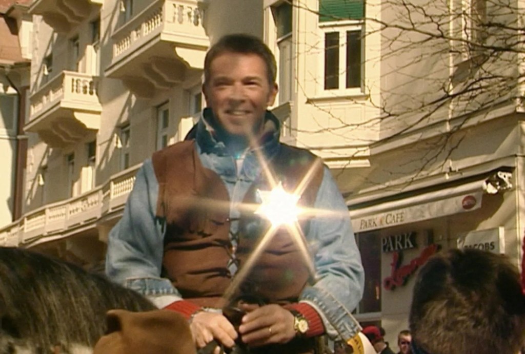 Mann auf einem Pferd, lächelnd und in Westernkleidung, mit Sonnenschein, vor einem Gebäude in der Stadt.