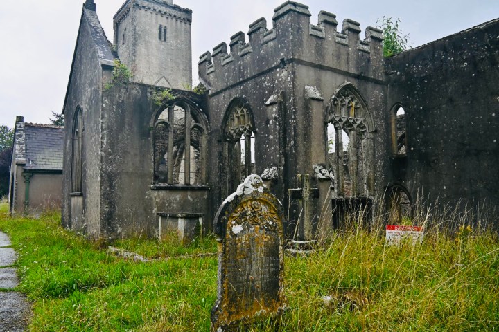 Ruinen einer alten Kirche mit einem verwitterten Grabstein im Vordergrund, umgeben von hohem Gras.