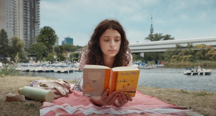 Ein Mädchen liegt auf einer Decke am Ufer eines Gewässers und liest ein Buch. Im Hintergrund sind Boote und moderne Gebäude zu sehen.