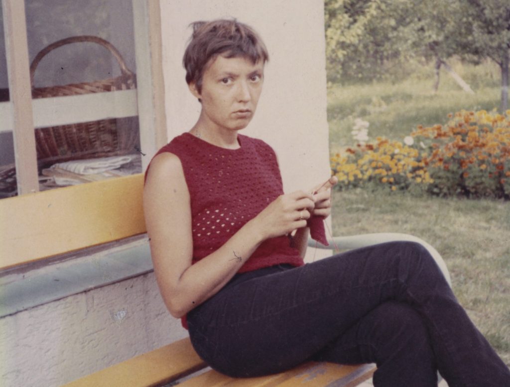 Eine Frau mit kurzen Haaren sitzt auf einer Bank im Freien, trägt ein rotes Oberteil und schaut ernst in die Kamera. Im Hintergrund sind Blumen und ein Fenster mit einem Korb sichtbar.